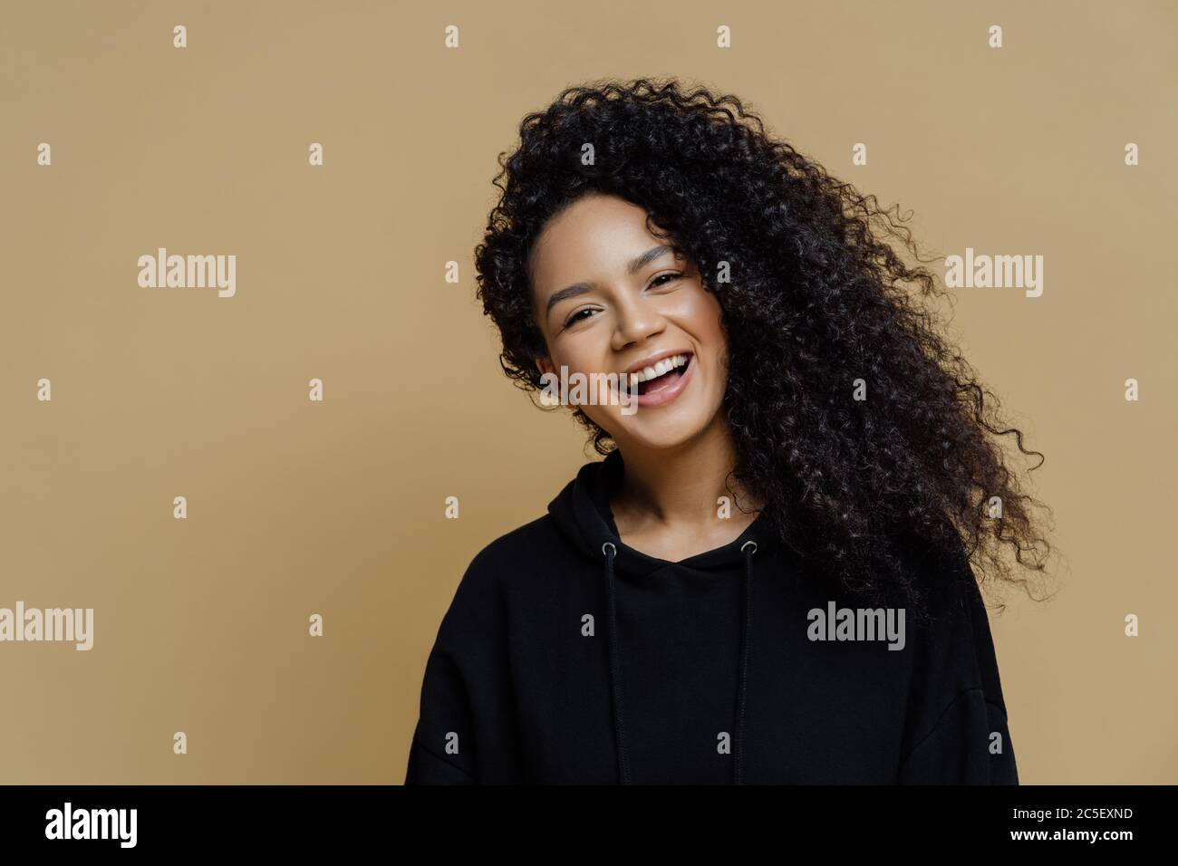 Felice positivo afroamericano donna con capelli ricci, inclina la testa e sorride ampiamente, vestito in casual felpa nera, isolato su sfondo beige Foto Stock