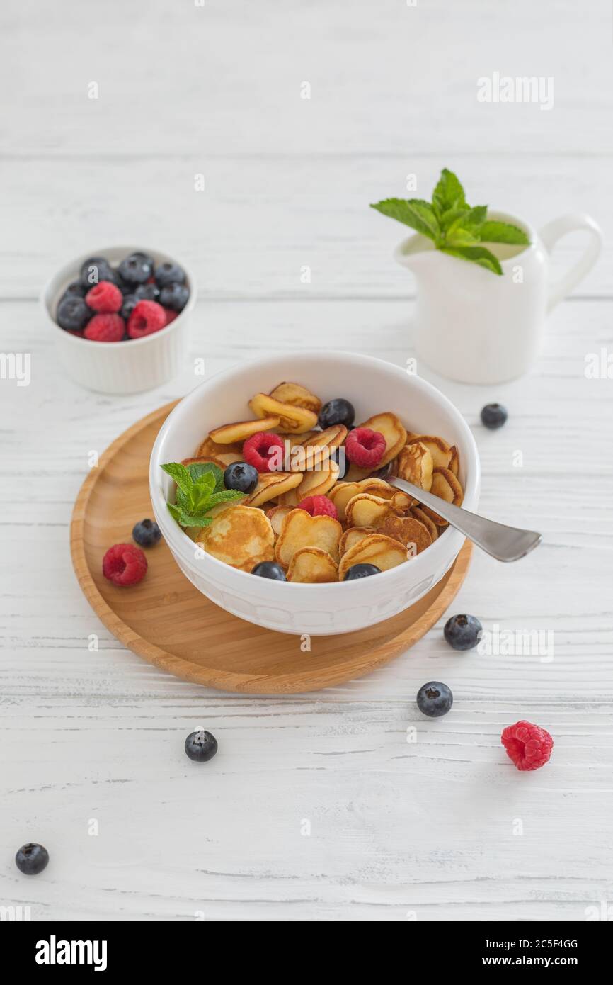 Prima colazione ciotola bianca con minuscoli cereali pancake con mirtilli, lamponi e menta su sfondo di legno Foto Stock