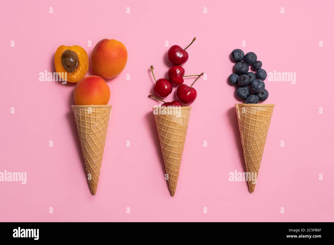 Vista dall'alto dei coni di cialde con frutta estiva (albicocche, mirtillo, ciliegia) su morbido sfondo rosa Foto Stock