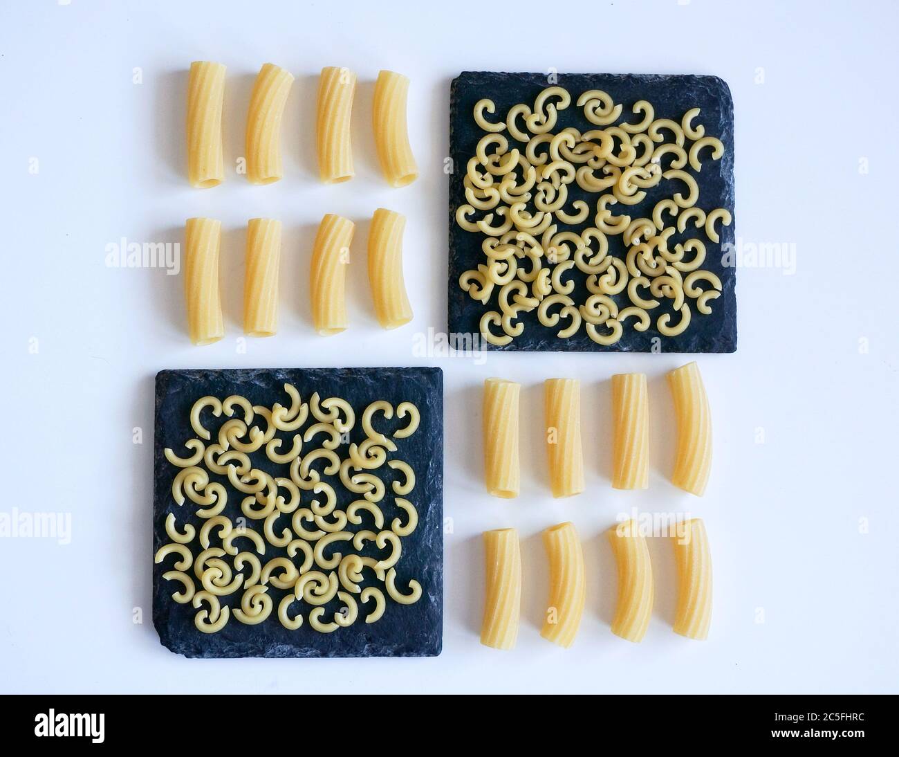 Diversi tipi di pasta cruda su coste quadrate di granito nero e sfondo bianco, concetto di lavorazione del cibo. Foto Stock