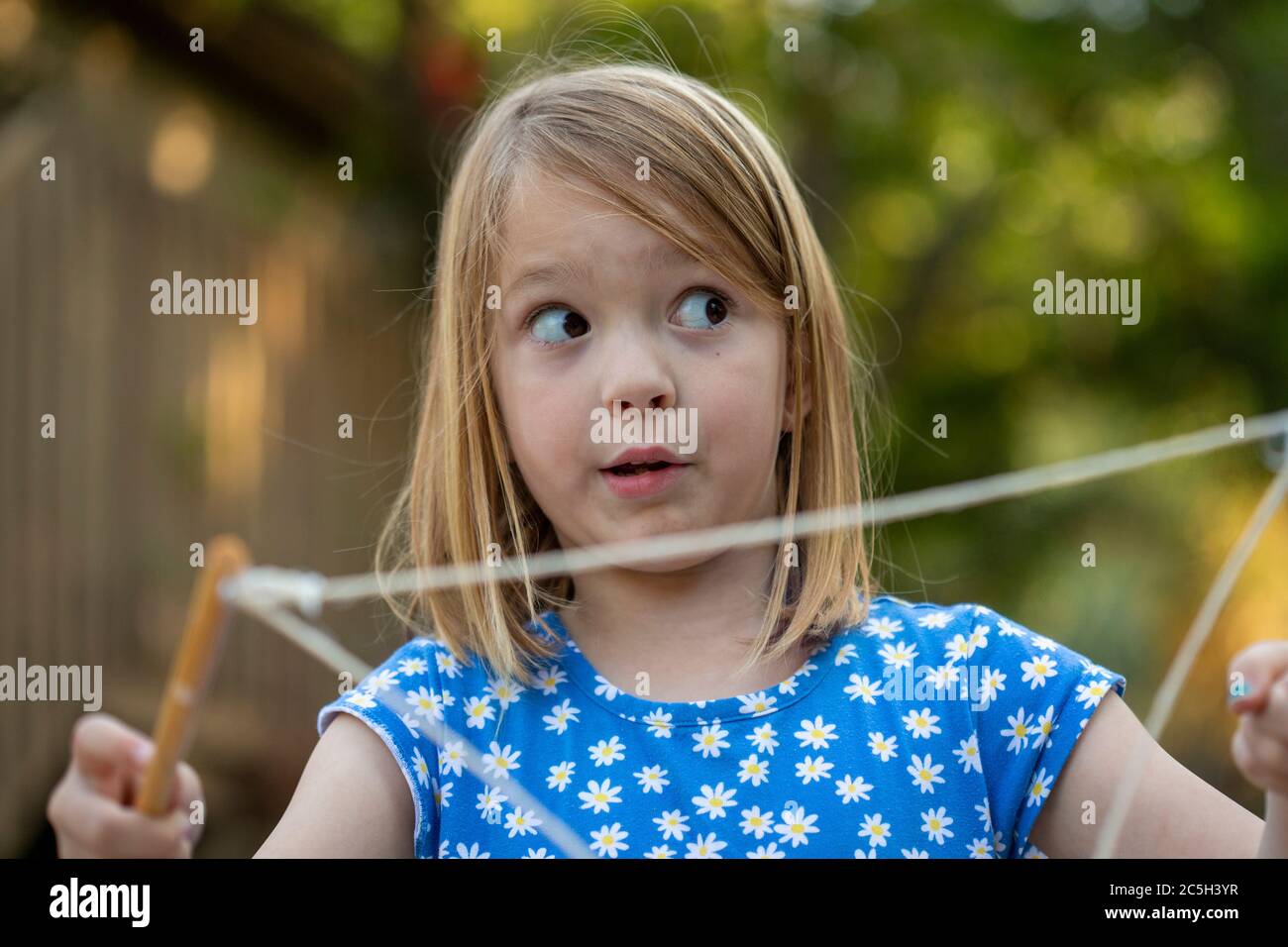 Giovane ragazza in un vestito blu e bianco che soffia grandi bolle con corda in un cortile posteriore Foto Stock