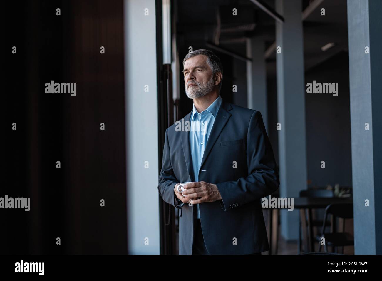 Uomo d'affari maturo che beve un caffè e che guarda fuori da una finestra della città da un edificio di ufficio. Foto Stock