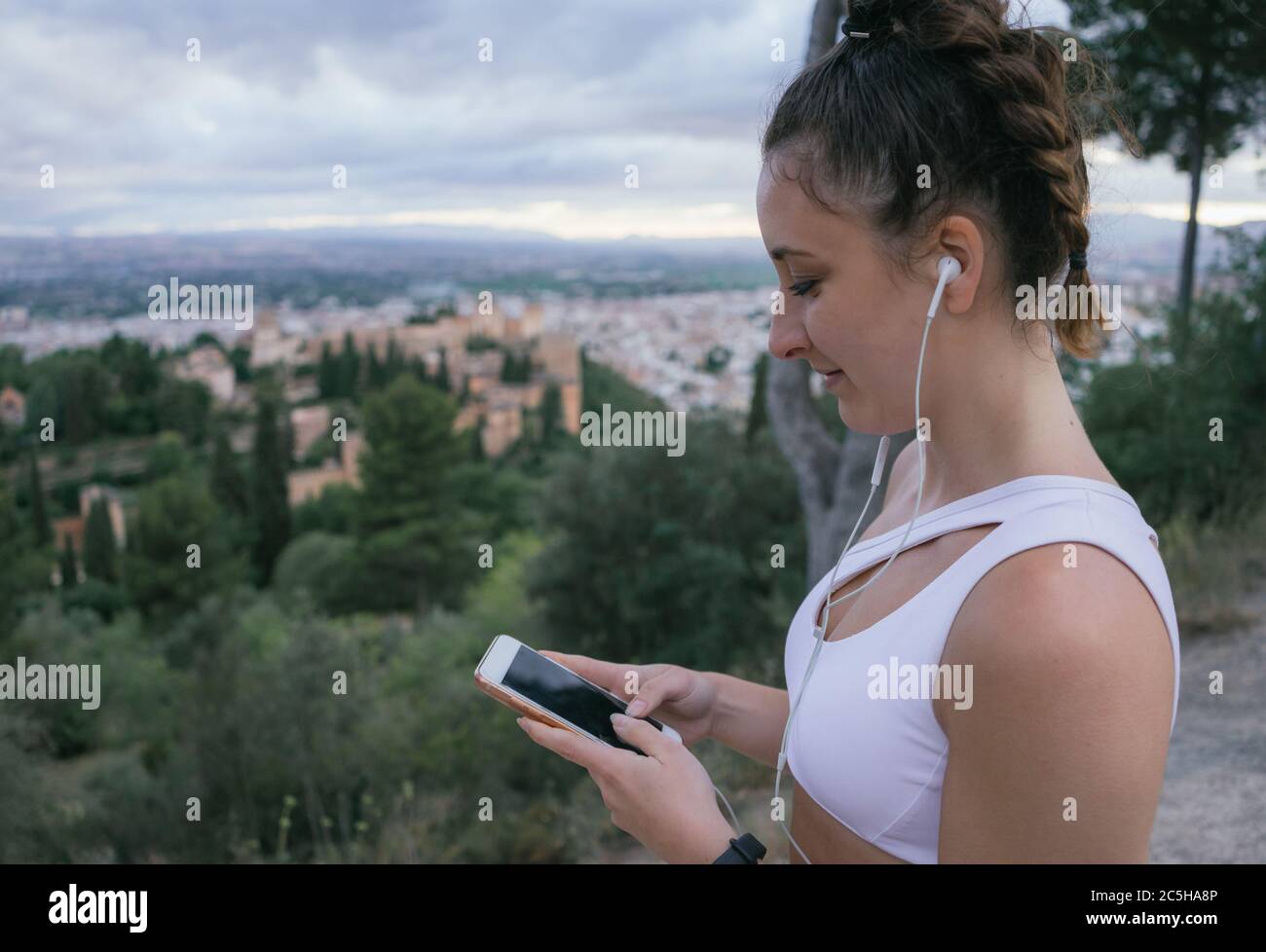 Immagine orizzontale di una ragazza felice con cuffie e telefono mentre riposa dal fare sport sopra la città dell'Alhambra in un pomeriggio estivo nuvoloso. Foto Stock Immagine orizzontale di una ragazza felice con cuffie e telefono mentre riposa dal fare sport sopra la città dell'Alhambra in un pomeriggio estivo nuvoloso. Foto Stock