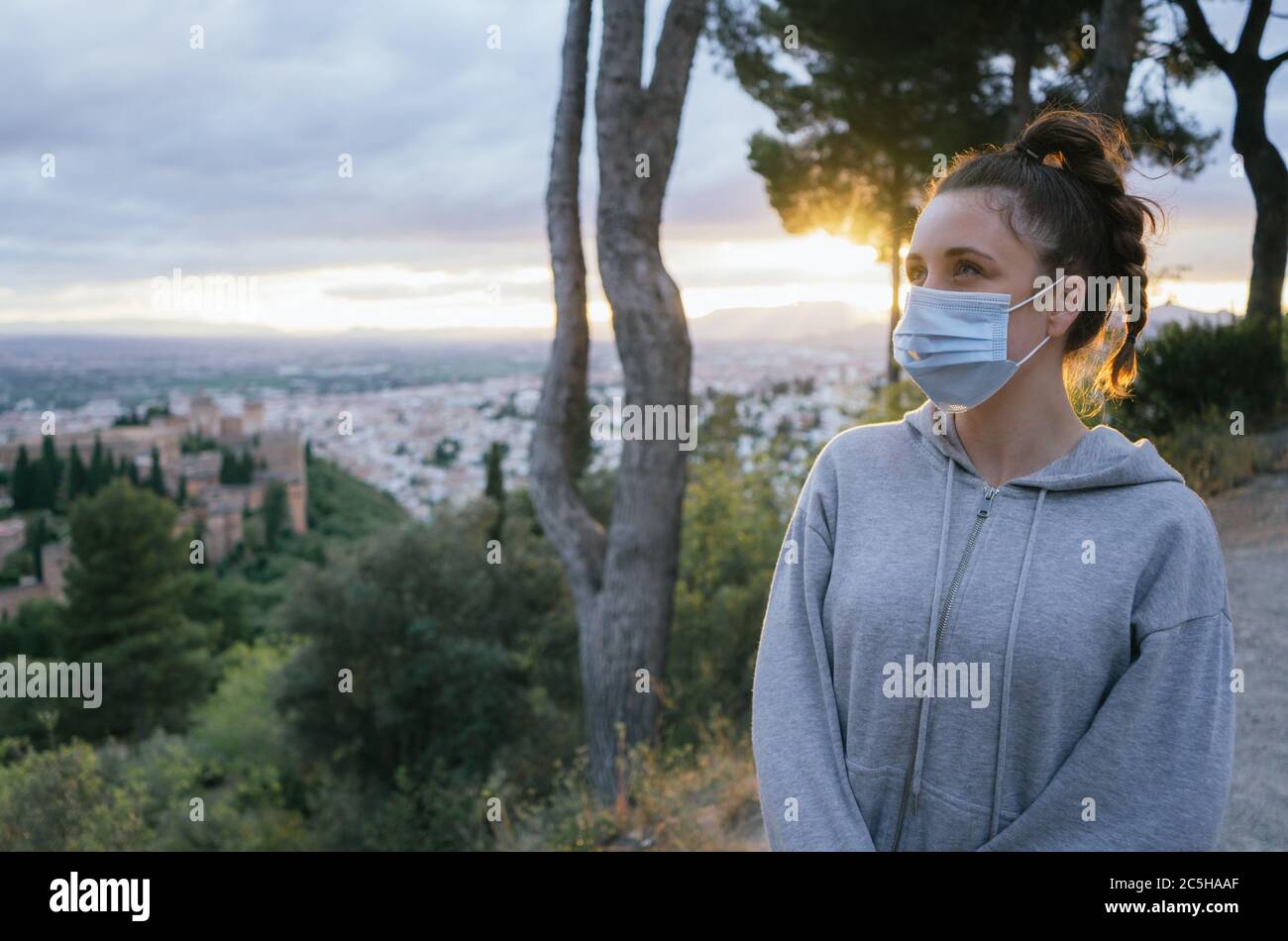 Giovane ragazza che guarda con una maschera dal covid, inquinamento con il sole in background, sopra la città dell'Alhambra, Spagna. Tramonto estivo. Foto Stock