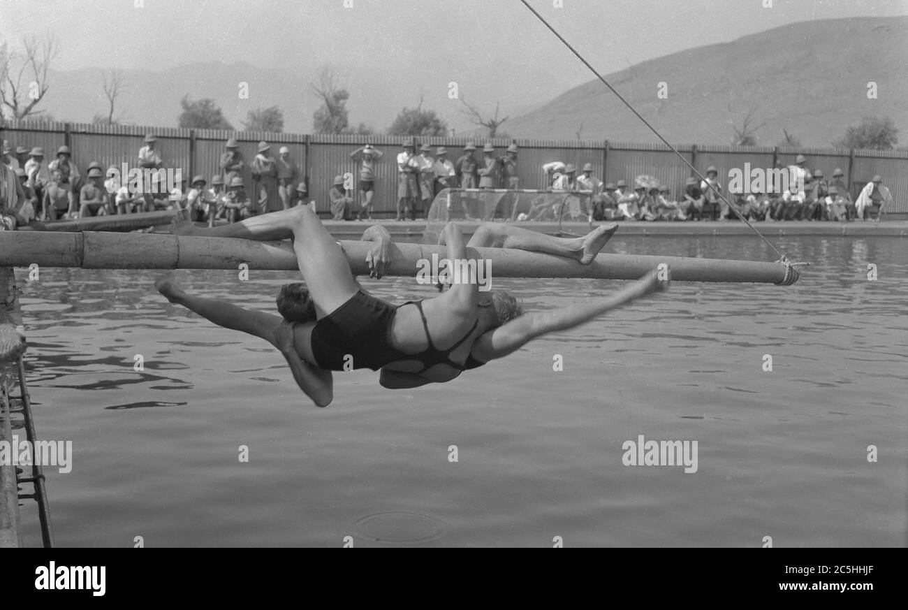 Circa, 1910, storico, due uomini nei costumi di bagno dell'epoca che si tiene su un palo di bandiera appeso sopra l'acqua in una grande piscina all'aperto, cercando di bussare o di combattere l'altro nell'acqua sottostante, forse Egitto. Simile in alcuni aspetti allo sport di wrestling ma sopra terra, l'attività richiede grande forza e agilità per rimanere sul palo. Foto Stock