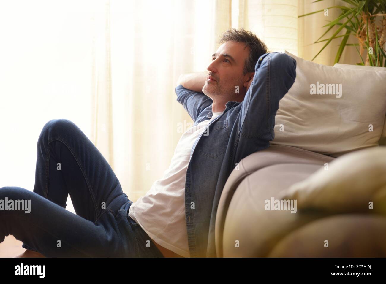 Uomo pendente seduto sul pavimento e appoggiato su un divano nel soggiorno di casa Foto Stock