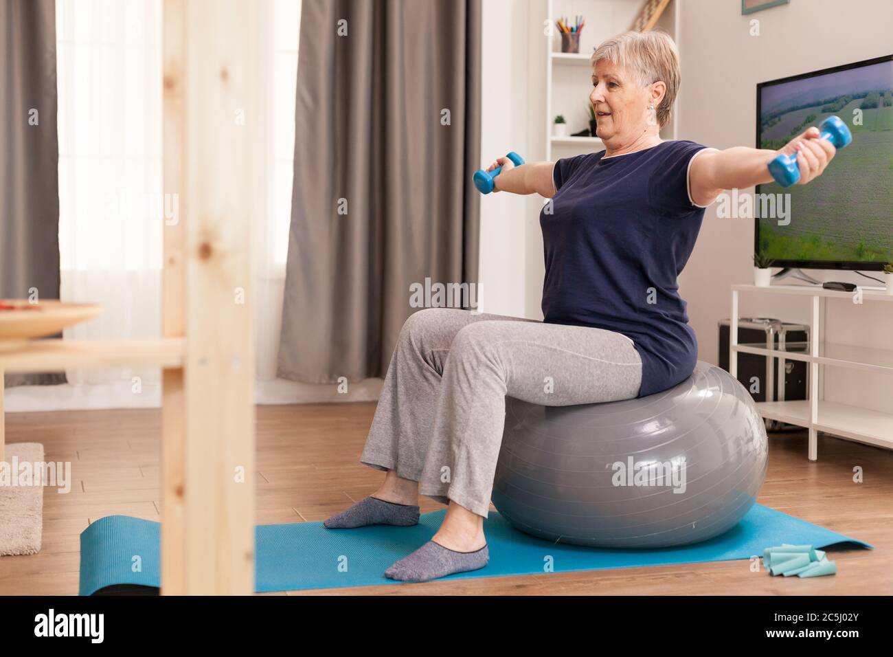 Vecchia casalinga che usa i dumbbells per il recupero fisico a casa. Anziano pensionato online internet esercizio di formazione a casa attività sportiva con banda di resistenza, palla svizzera all'età anziana di pensionamento Foto Stock