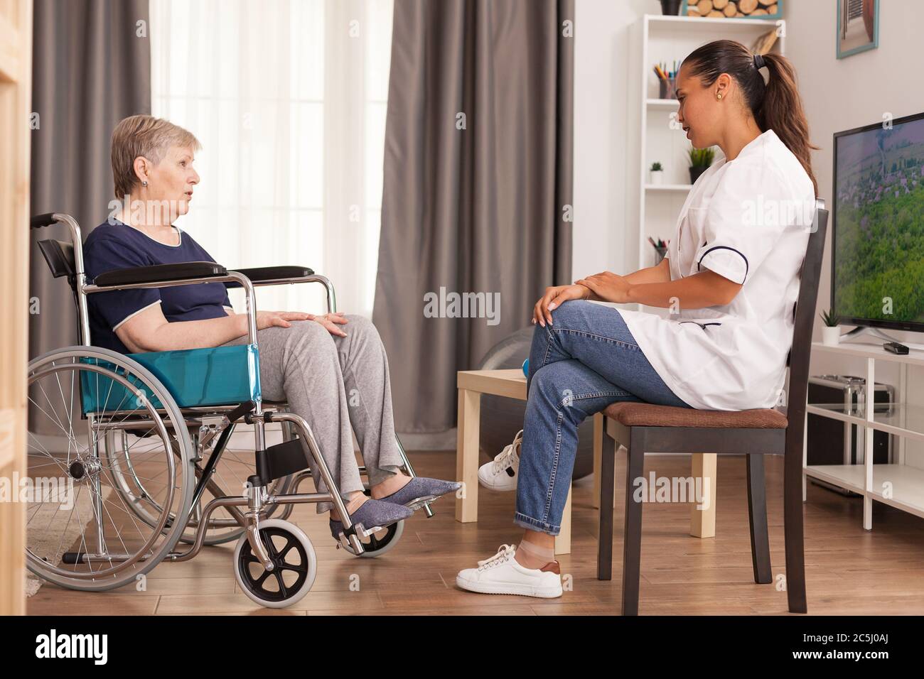 Vecchia signora malata che parla con l'assistenza sociale seduta in una sedia a rotelle nel suo appartamento. Disabili anziani ricoverati infermiere professionista di aiuto, cura casa di riposo e riabilitazione Foto Stock