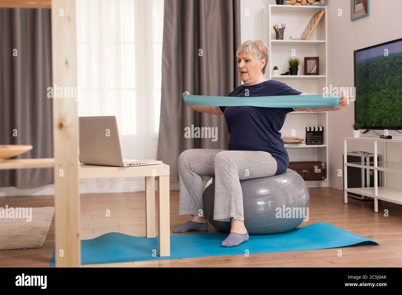 Donna anziana che si allena a casa davanti al computer portatile. Anziano pensionato online internet esercizio di formazione a casa attività sportiva con manubri, banda di resistenza, palla svizzera all'età di anziano pensionamento Foto Stock