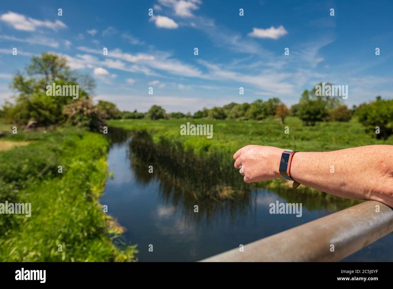 Donna con braccia ripiegate vista su un piccolo ponte fluviale nella campagna inglese. Lei indossa un orologio fitness e le sue braccia sono abbronzate. Foto Stock