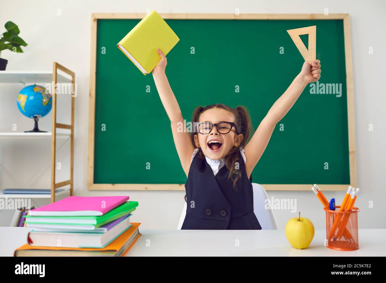 Ritorno a scuola. Un bambino piccolo e felice ha alzato le mani sullo sfondo di una lavagna verde in una classe. Foto Stock