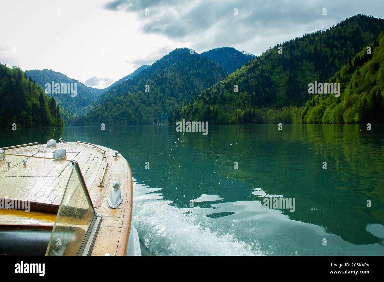 Barca retrò a vela sul lago di Ritsa tra le montagne. Le onde divergono dal lato dell'imbarcazione. Maestose montagne incorniciano il lago blu di Ritsa. PE Foto Stock