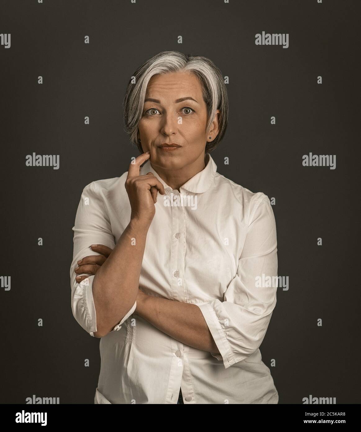 La donna penosa tocca il viso con la mano mentre guarda la fotocamera. Donna matura in camicia bianca isolata su sfondo grigio. Concetto di tempesta cerebrale. Oscurata Foto Stock