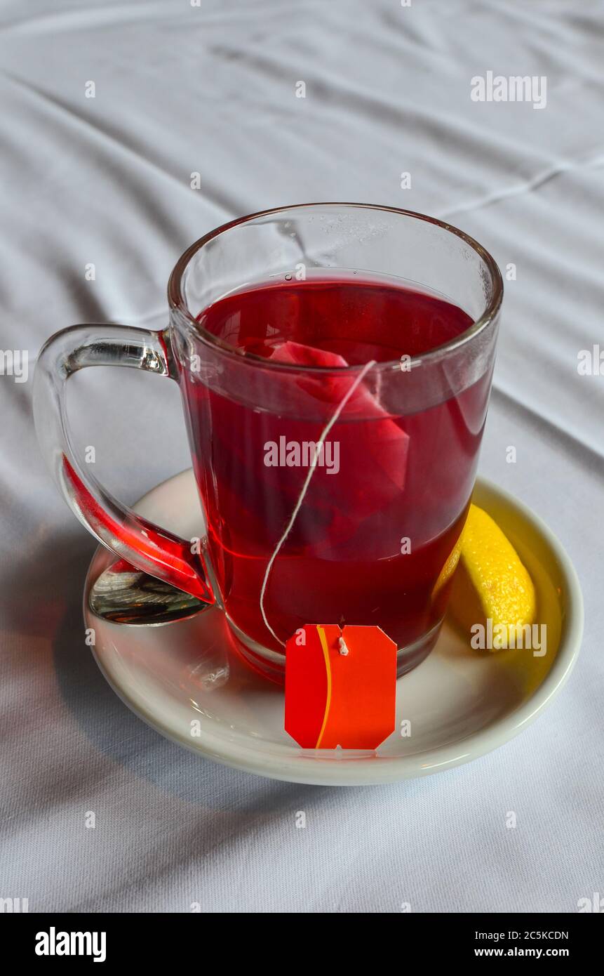 Sacchetto filtrante di tè ibisco in un bicchiere su piattino bianco in ceramica con fetta di limone Foto Stock