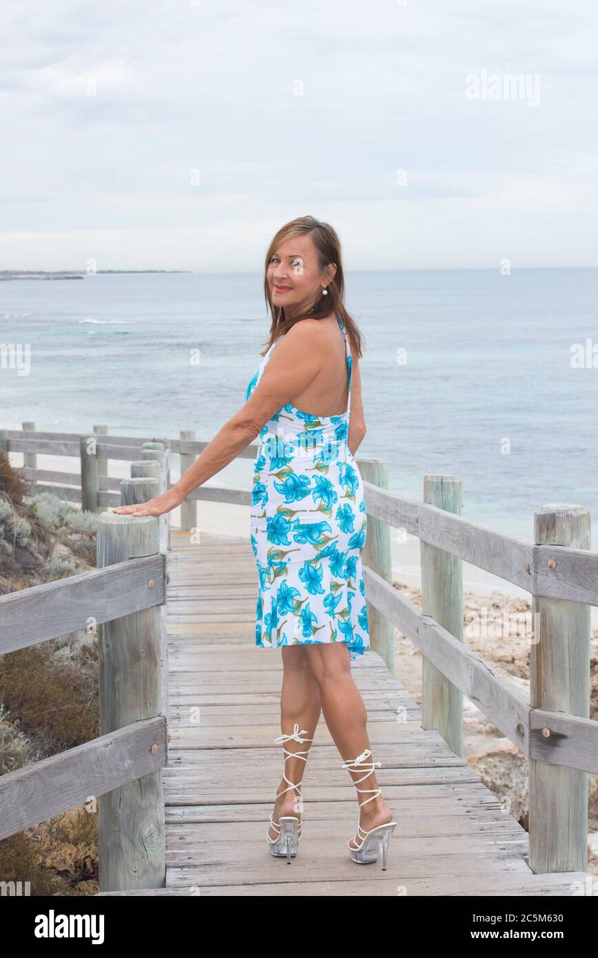 Ritratto sicuro bella donna matura che posa felice sorridente e rilassato in piedi in alto scarpe tallone esterno a costa, con oceano e orizzonte come blu Foto Stock