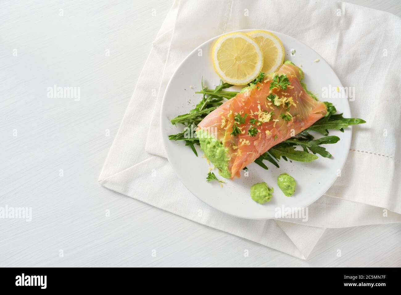 Rotolo di salmone con purea di piselli ripieno su insalata di razzo con fette di limone e guarnitura di erbe su un tavolo bianco, sano dimagrimento con carb basso, spazio di copia, hi Foto Stock