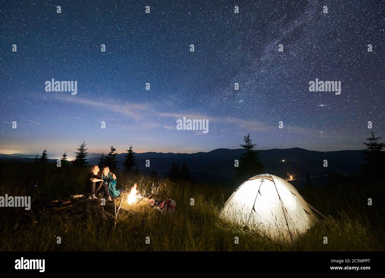 Amici escursionisti seduti su panchina fatto di tronchi, guardando insieme incredibile cielo stellato in montagne accanto a tenda illuminata, cresta di montagna sullo sfondo. Concetto di escursioni, campeggio notturno, relazioni Foto Stock