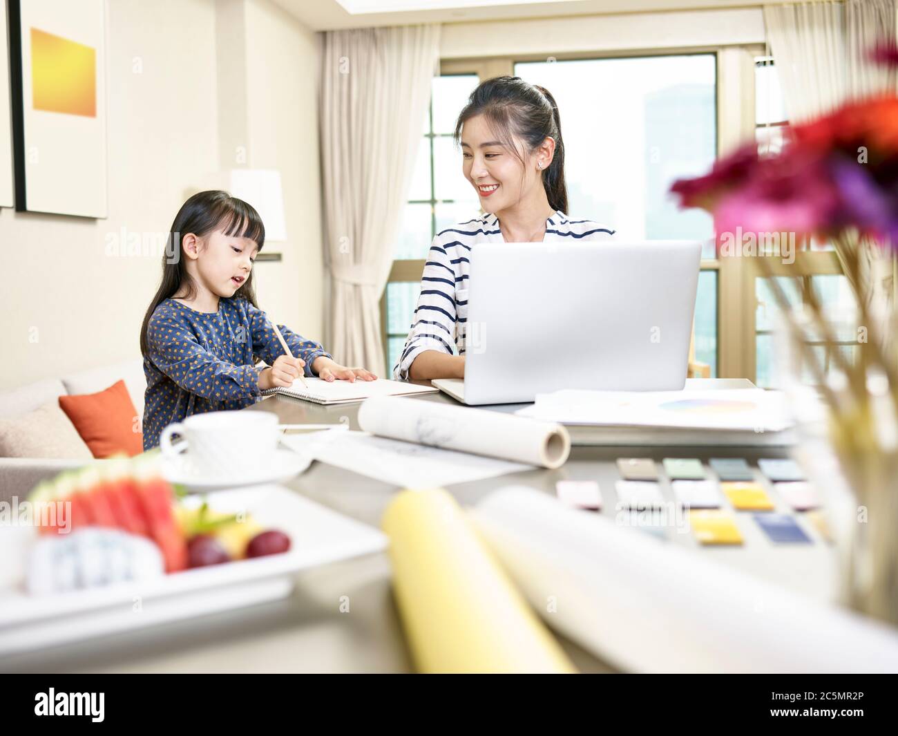 giovane designer asiatica madre che lavora da casa utilizzando un computer portatile mentre si prende cura di figlia (grafica in background alterato digitalmente) Foto Stock