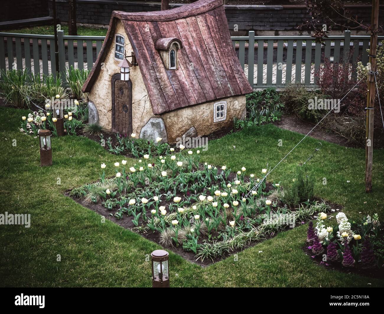 Russia, Sochi 14.03.2020. Una piccola casa da favola sorge su un prato verde con un letto fiorito con tulipani lanterne basse e una recinzione dietro Foto Stock