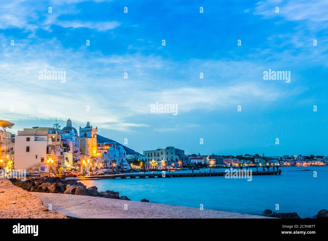 Vista sull'isola d'Ischia, con edifici e molo illuminati al crepuscolo con persone sullo sfondo che si godono la serata. Foto Stock
