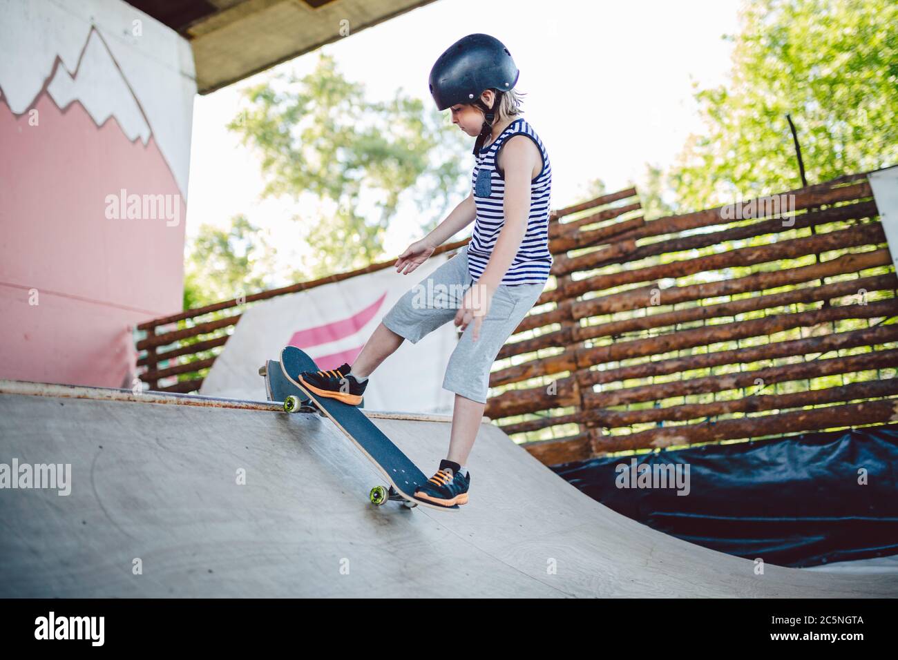 Caucasico ragazzo in un casco fa trucchi su uno skateboard su un parco giochi per skateboard all'esterno. Uno skateboarder per bambini fa una rampa di mezzi pipe su una Foto Stock