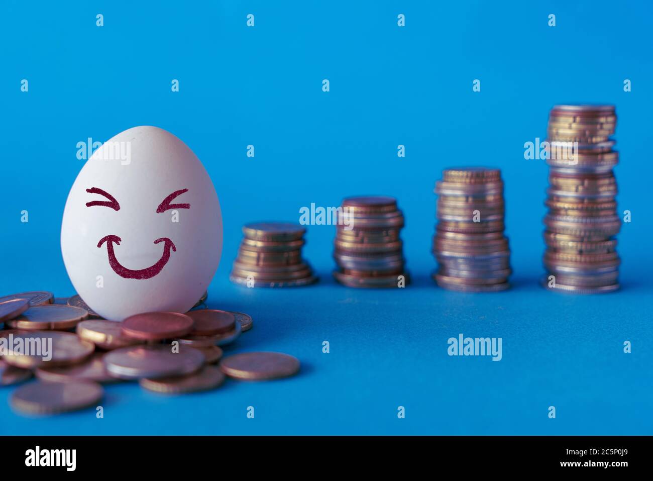 Facce sulle uova, concetto di investimento. Felice investitore sorridendo di fronte al suo crescente denaro. Sfondo blu con pile di monete Foto Stock