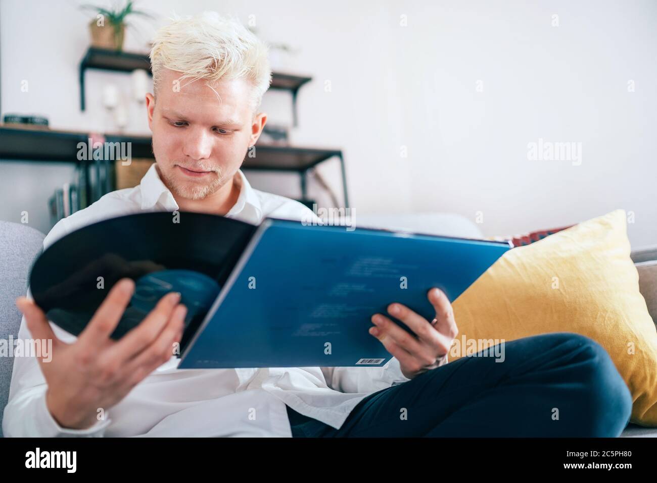 Ritratto di un giovane seduto in un comodo divano nel soggiorno che prende 12 pollici vinile LP dalla manica. Immagine del concetto di valore del suono analogico vintage Foto Stock