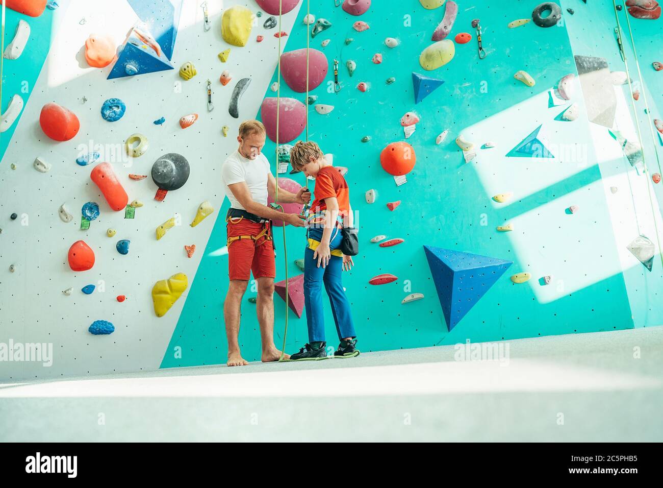 Padre che aiuta il figlio adolescente e controlla l'imbracatura da arrampicata vicino alla parete da arrampicata interna. Ragazzo che si prepara per un'arrampicata. Felice idea geniting imag Foto Stock