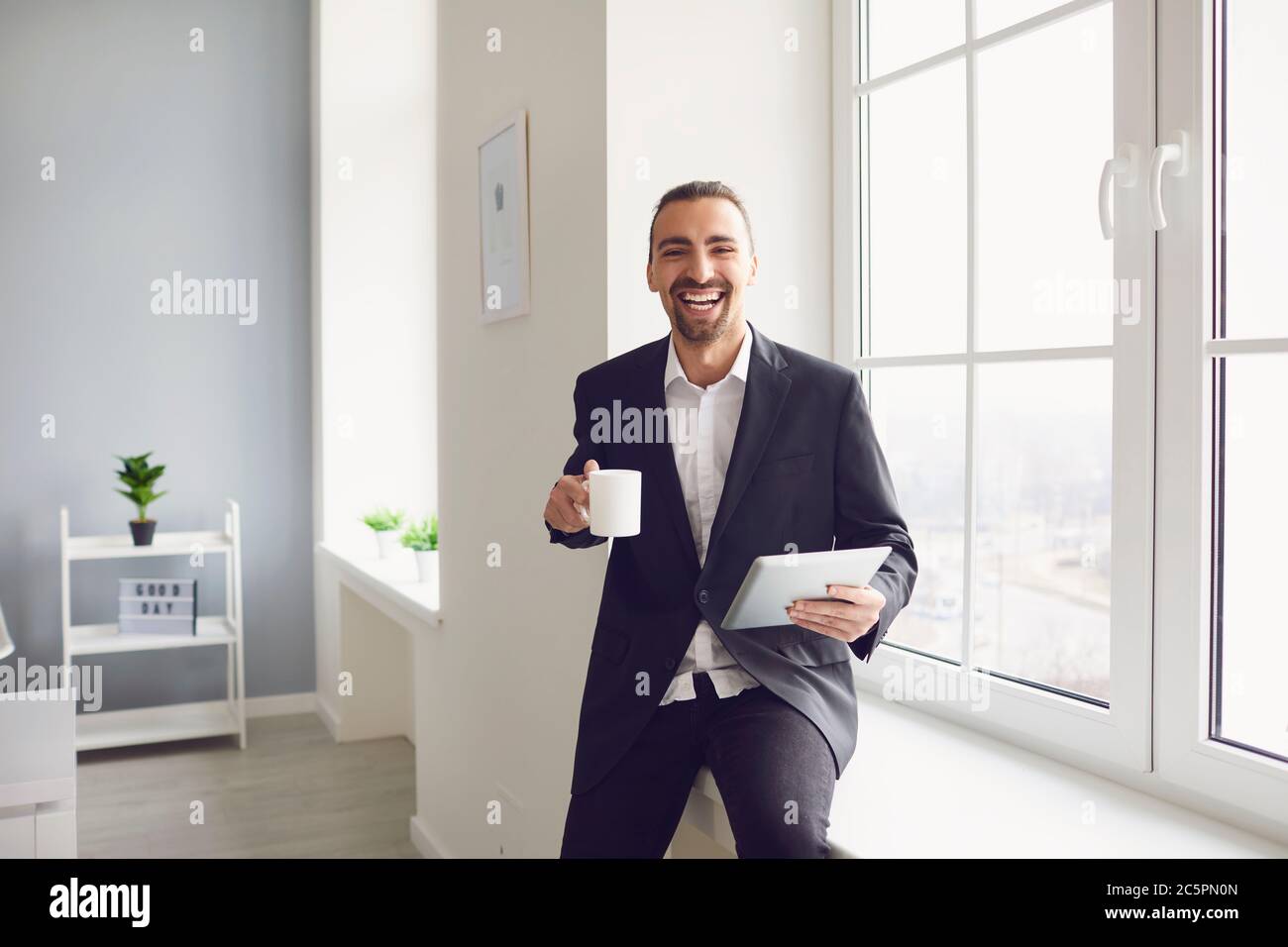 Un uomo in una giacca nera con una tazza di caffè in mano sorride mentre sta in piedi accanto alla finestra in ufficio Foto Stock