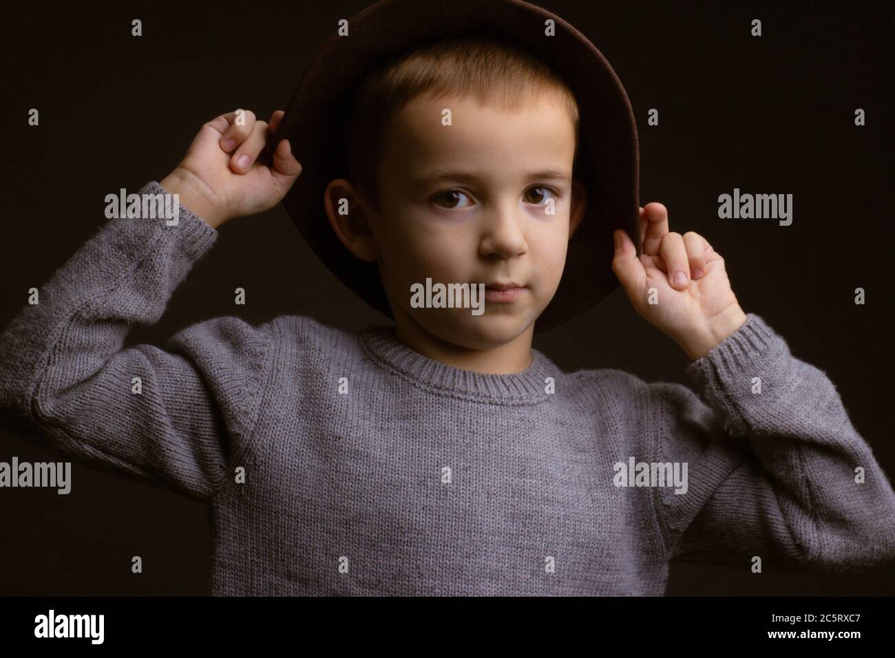 Studio ritratto di un ragazzo in un maglione grigio, su sfondo nero, che tiene un cappello marrone accanto alla sposa Foto Stock