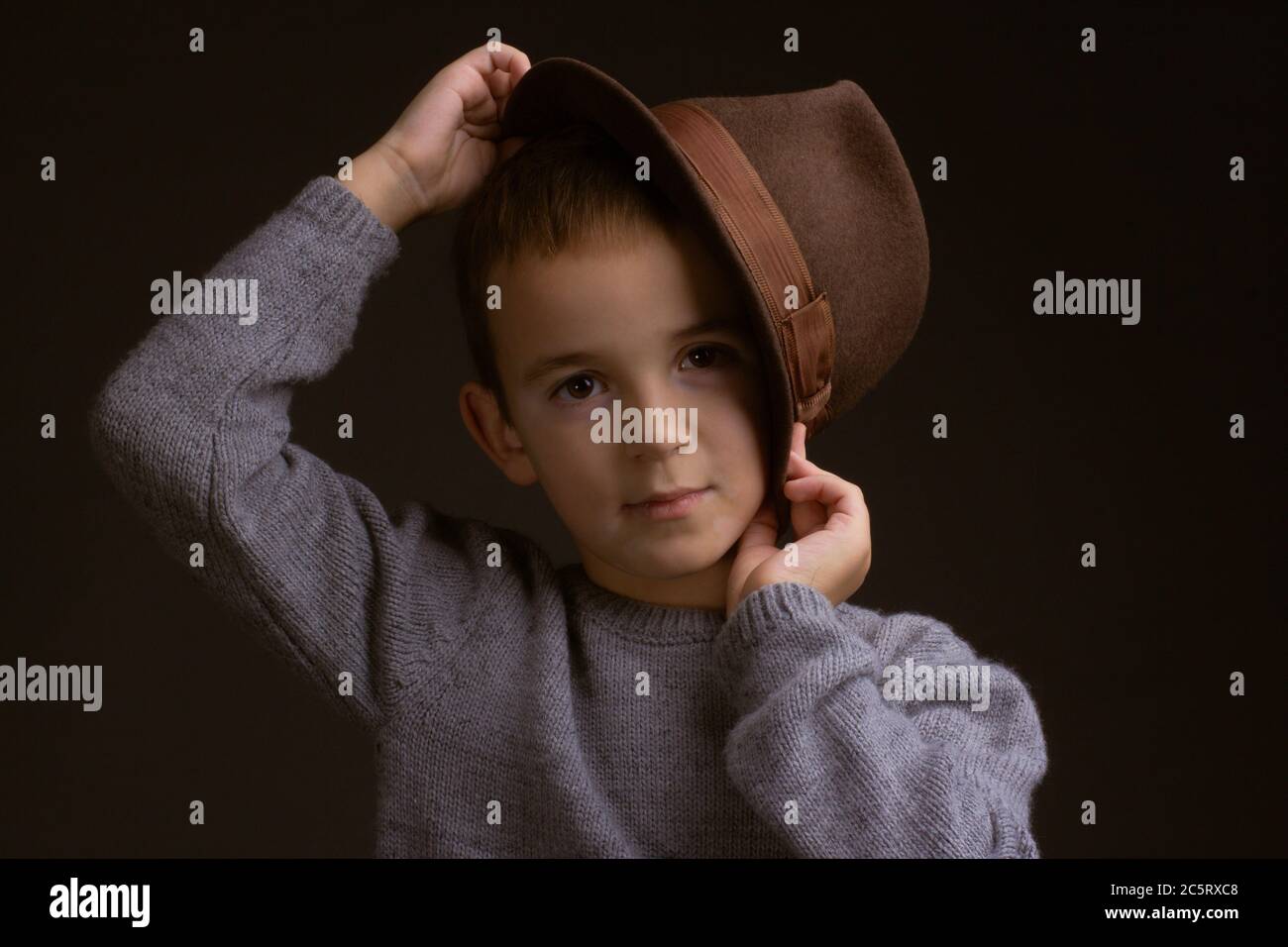 Studio ritratto di un ragazzo in un maglione grigio, su sfondo nero, che tiene un cappello marrone accanto alla sposa Foto Stock