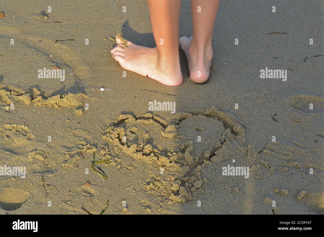 Disegno di un cuore nella sabbia e piedi di un bambino in piedi dietro Foto Stock