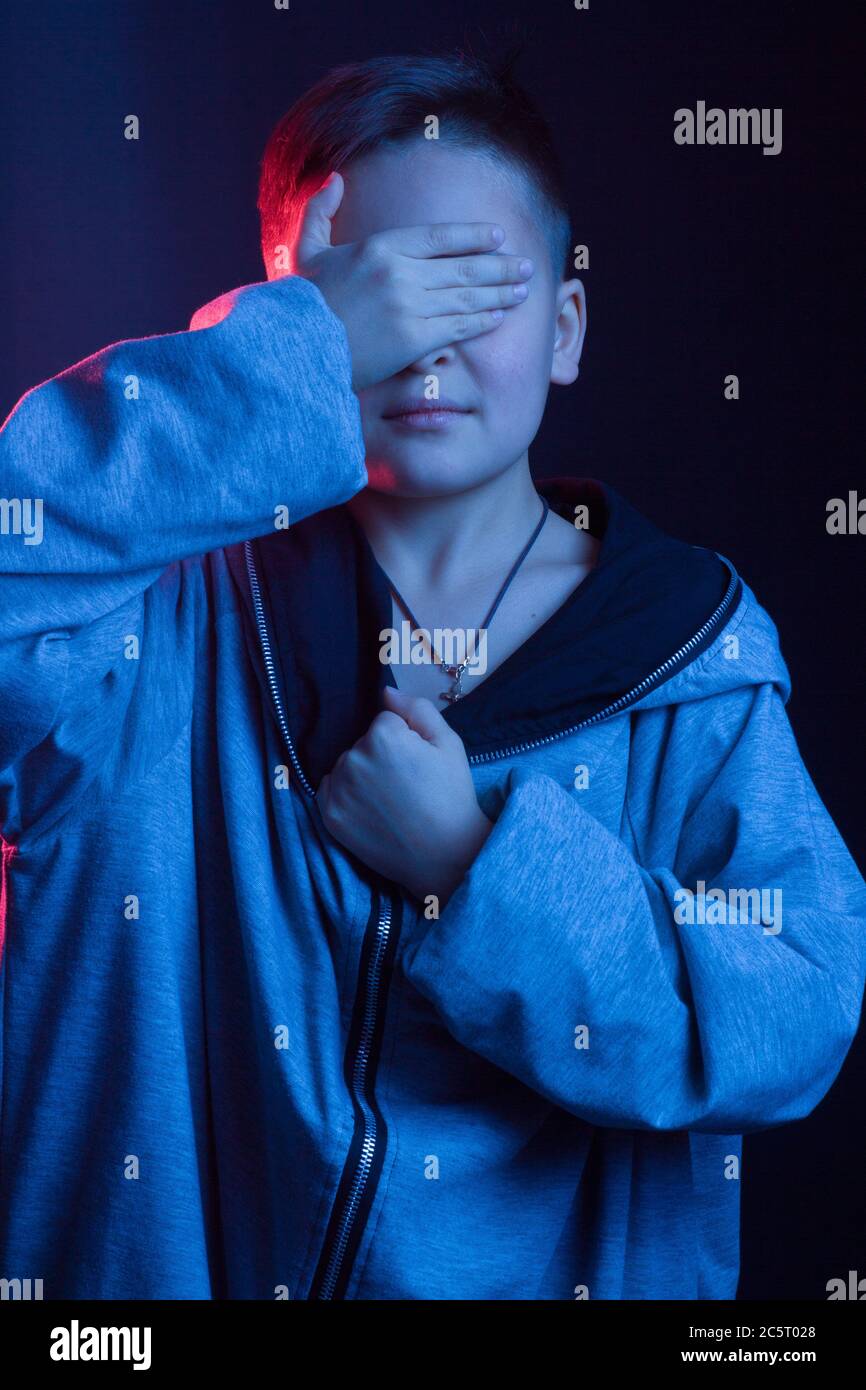 Ritratto studio di un ragazzo dai capelli scuri, su sfondo scuro, in una giacca con cappuccio. Colore chiaro: Rosso e blu. Ricopre il viso con la mano Foto Stock