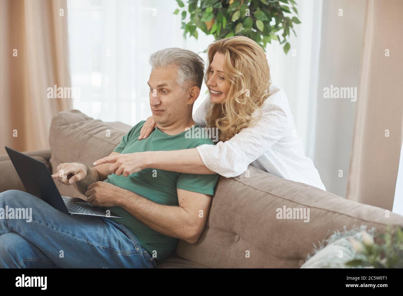 Ritratto laterale di un uomo maturo dai capelli bianchi che usa un computer portatile mentre si rilassa sul divano con una moglie sorridente che lo abbraccia da dietro e che punta verso lo schermo, Foto Stock