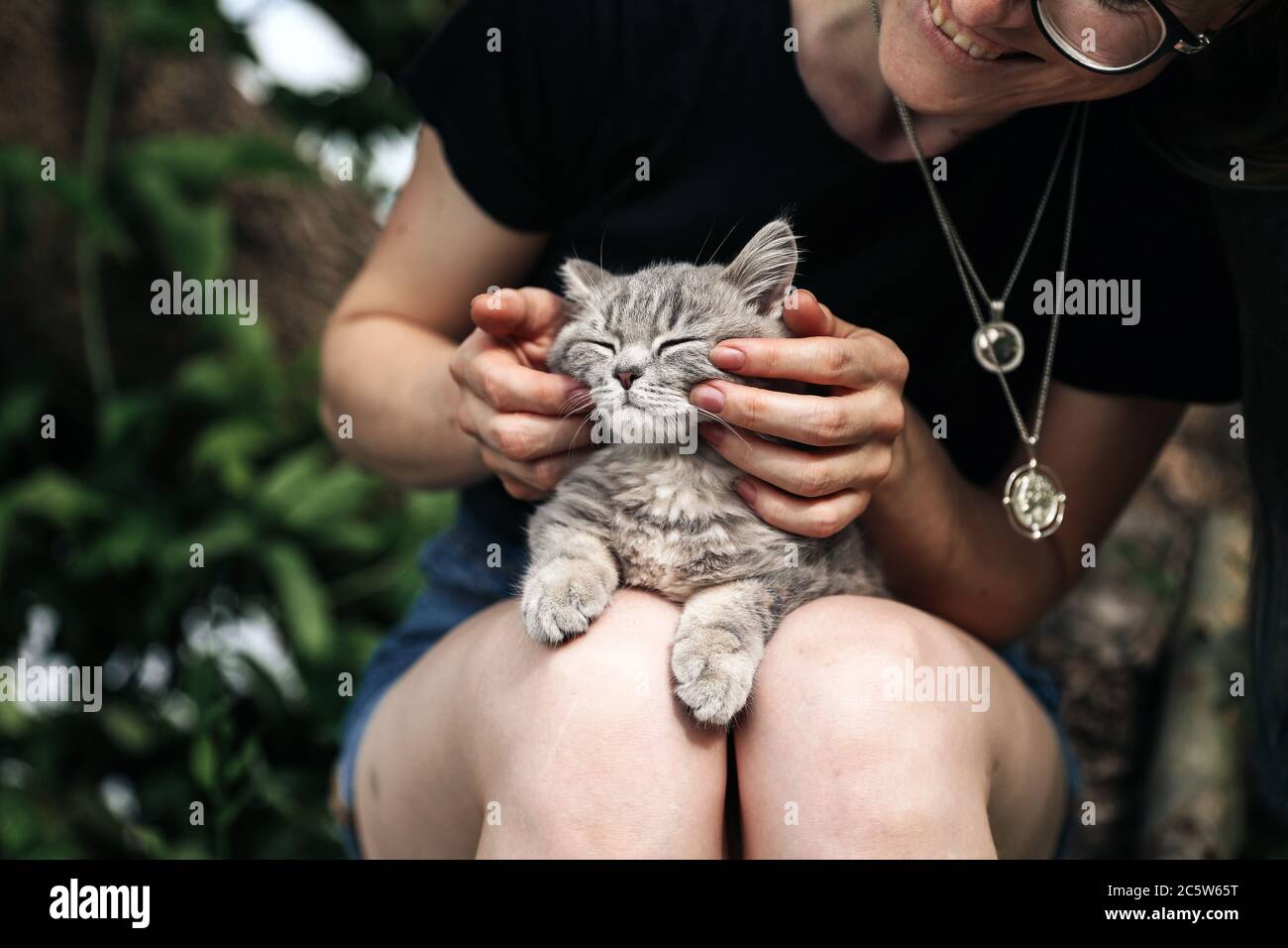 Una giovane donna tiene un gattino scozzese dritto all'aperto. Il gatto si diverte e si fa con gli occhi chiusi e sembra divertente. Primo piano, luce naturale Foto Stock