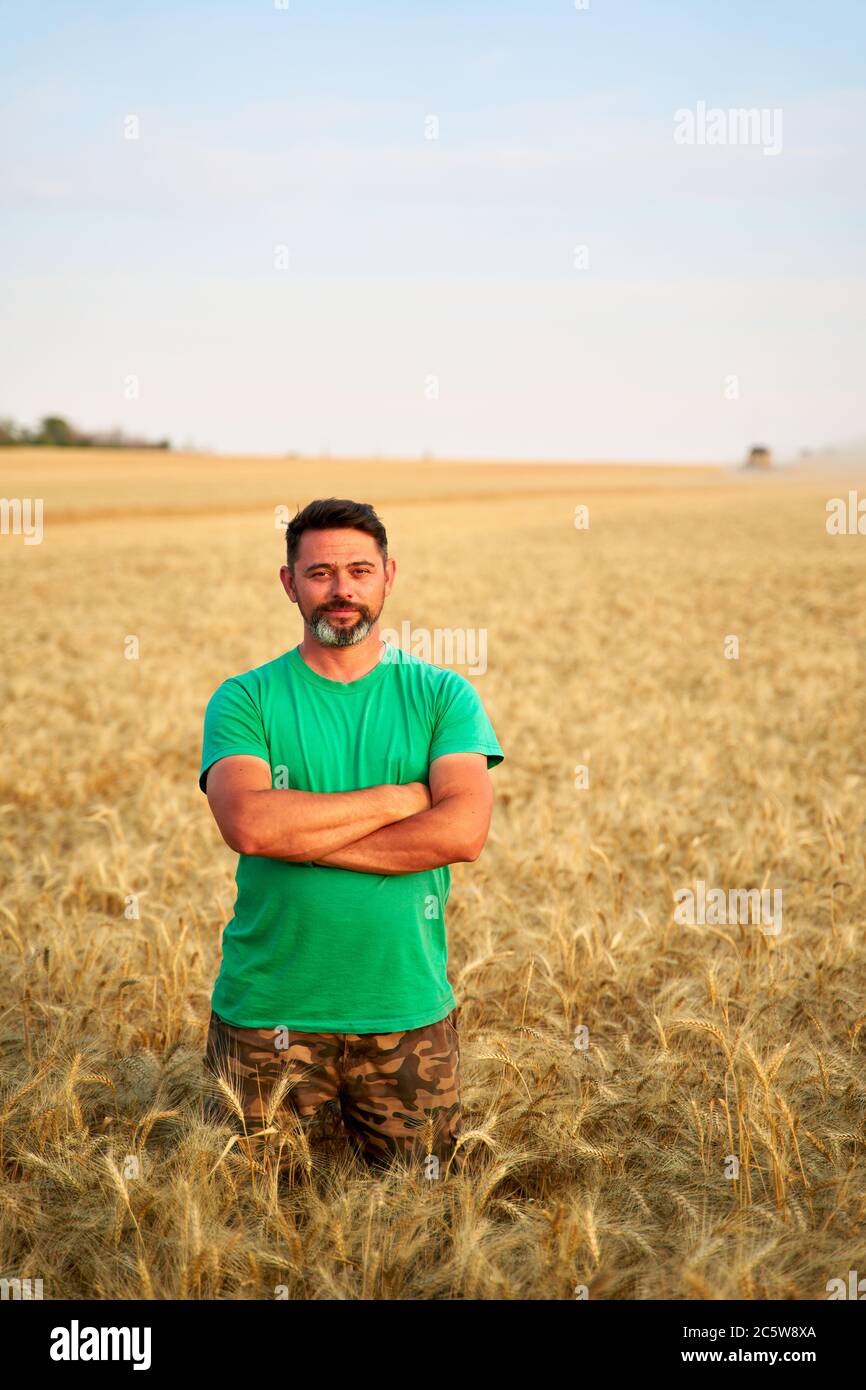 Agricoltore felice in piedi orgogliosamente in campo di grano con le braccia incrociate sul petto. Agronomo che indossa l'uniforme aziendale, guardando la macchina fotografica su terreni agricoli. Ricco Foto Stock