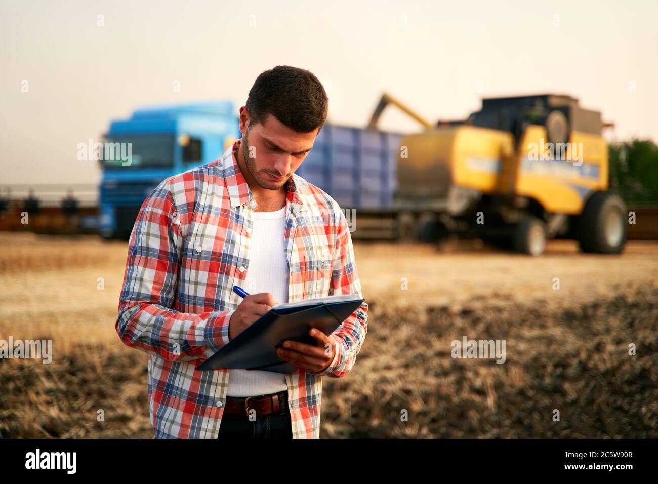 L'agricoltore controlla il caricamento del frumento dalla trebbiatrice al dumper per granella. Conducente che tiene appunti, tiene appunti, conta carichi. Lo spedizioniere compila la spedizione Foto Stock
