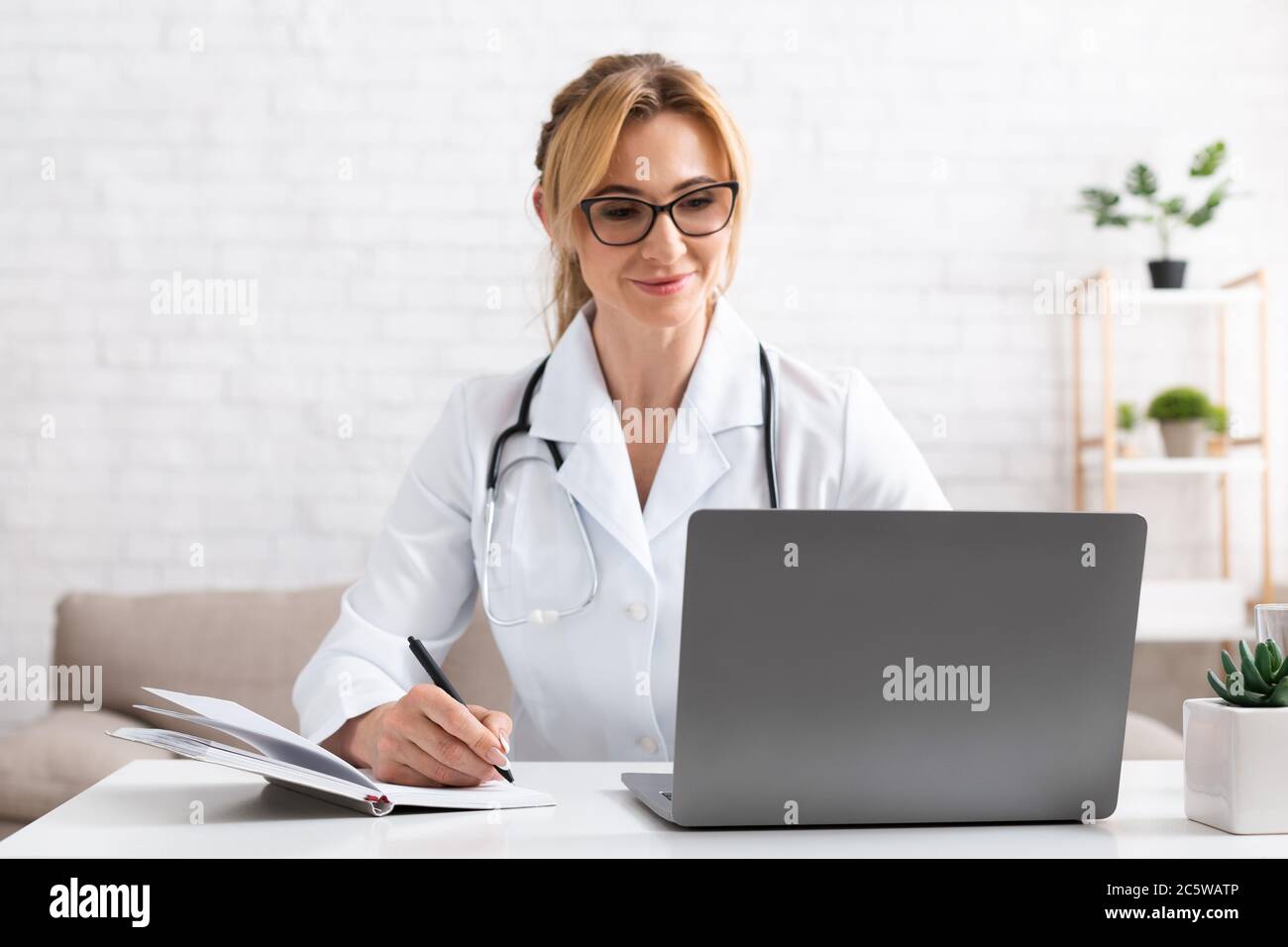 Il paziente parla con il medico utilizzando la videochiamata sul computer portatile. Donna in camice bianco prendere appunti in notebook Foto Stock