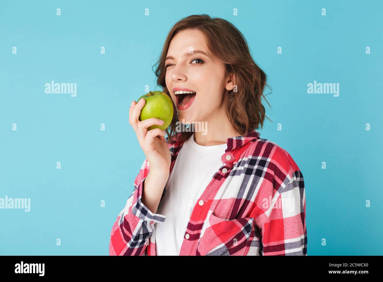Bella signora in camicia plaid mangiare mela verde e felicemente winking guardando in macchina fotografica su sfondo blu colorato Foto Stock