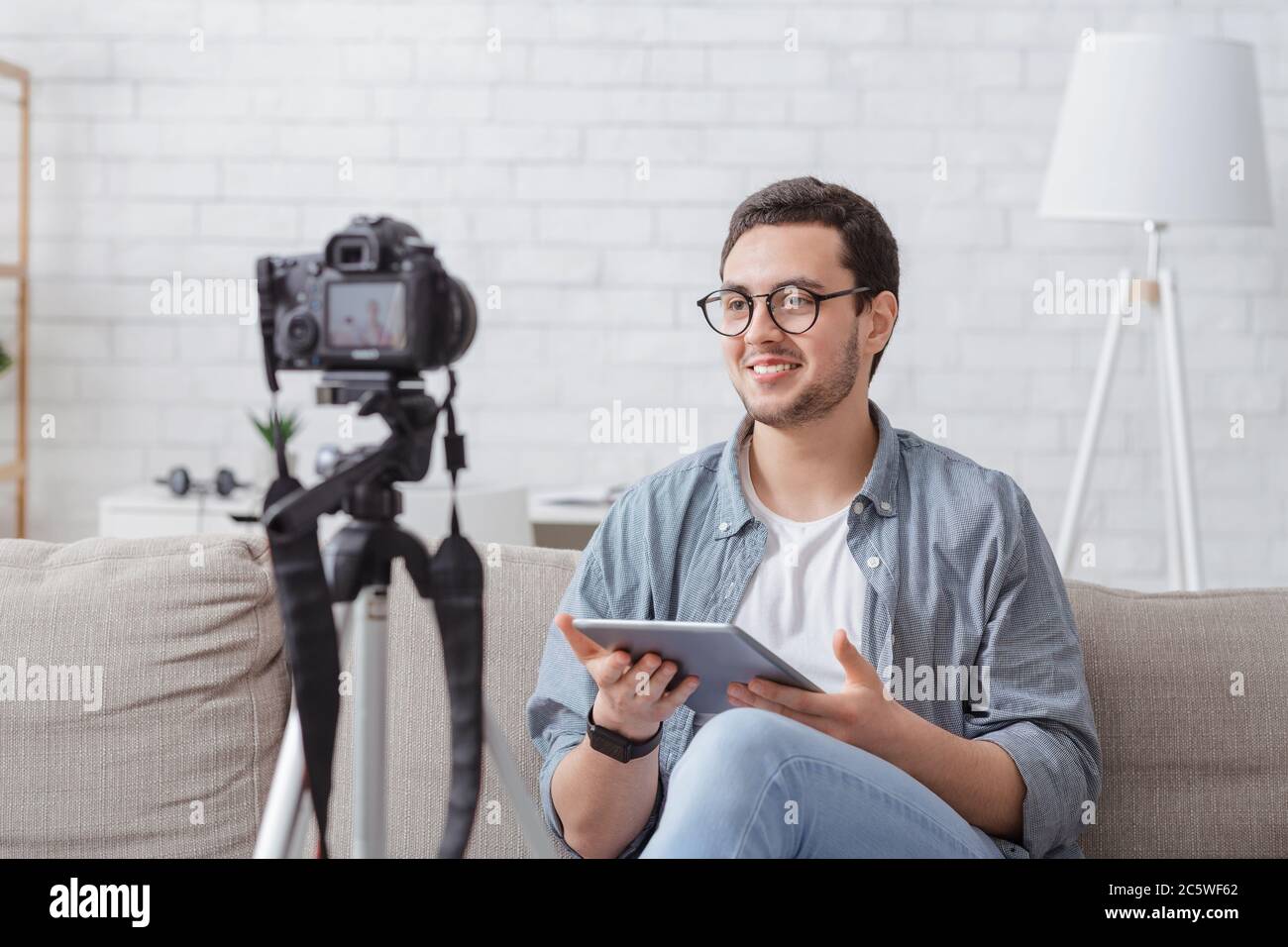 Video blogger sul posto di lavoro a casa. Sorridente giovane in occhiali con tablet parla alla fotocamera Foto Stock
