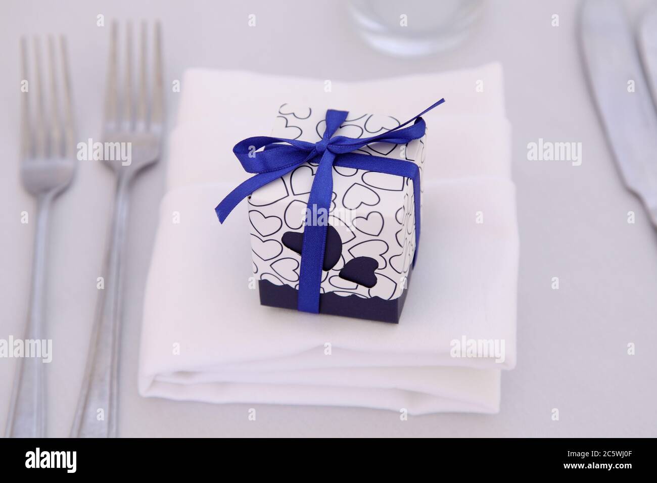 Piccolo regalo avvolto in un nastro blu con arco sul tavolo da pranzo con posate e tovaglioli. Favori di nozze o favori di nozze su sfondo bianco. Foto Stock