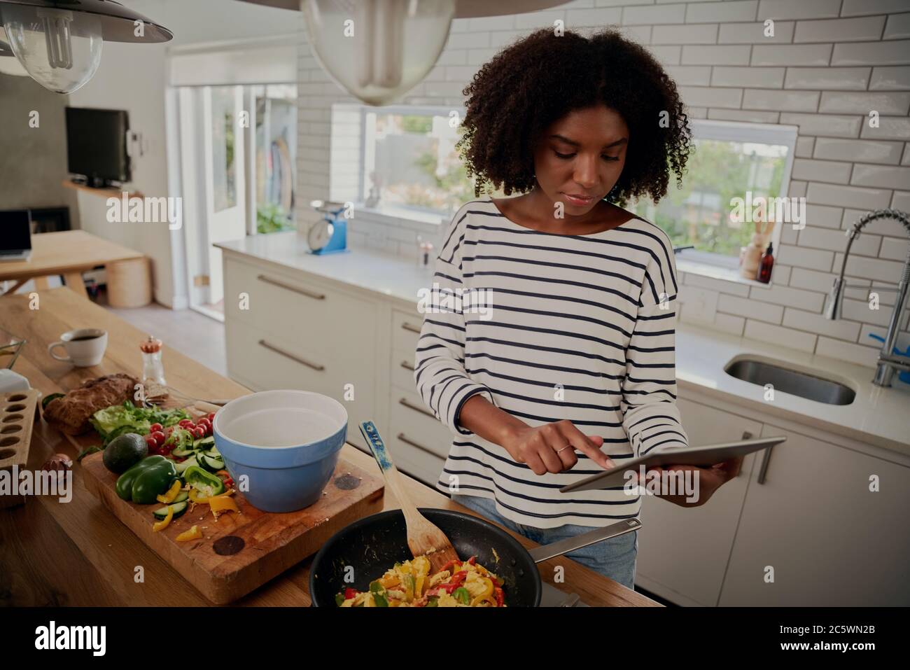 Giovane donna africana che guarda la ricetta in un tablet digitale mentre si cucina il pranzo in cucina moderna Foto Stock