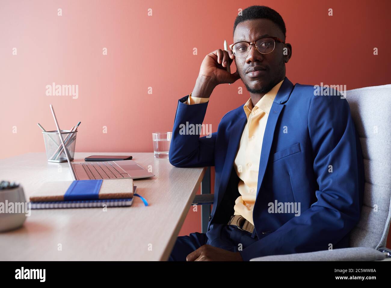 Ritratto di giovane uomo d'affari afroamericano che guarda via mentre si posa sul posto di lavoro in interni ufficio contro parete rossa, spazio copia Foto Stock