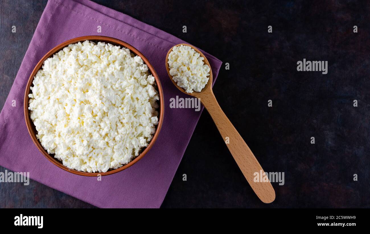 Ricotta il formaggio e cucchiaio di legno su sfondo scuro. Formaggio fresco casolare in una ciotola. Piatto di argilla con formaggio morbido su un tovagliolo di lino. Vista dall'alto. Copia sp Foto Stock