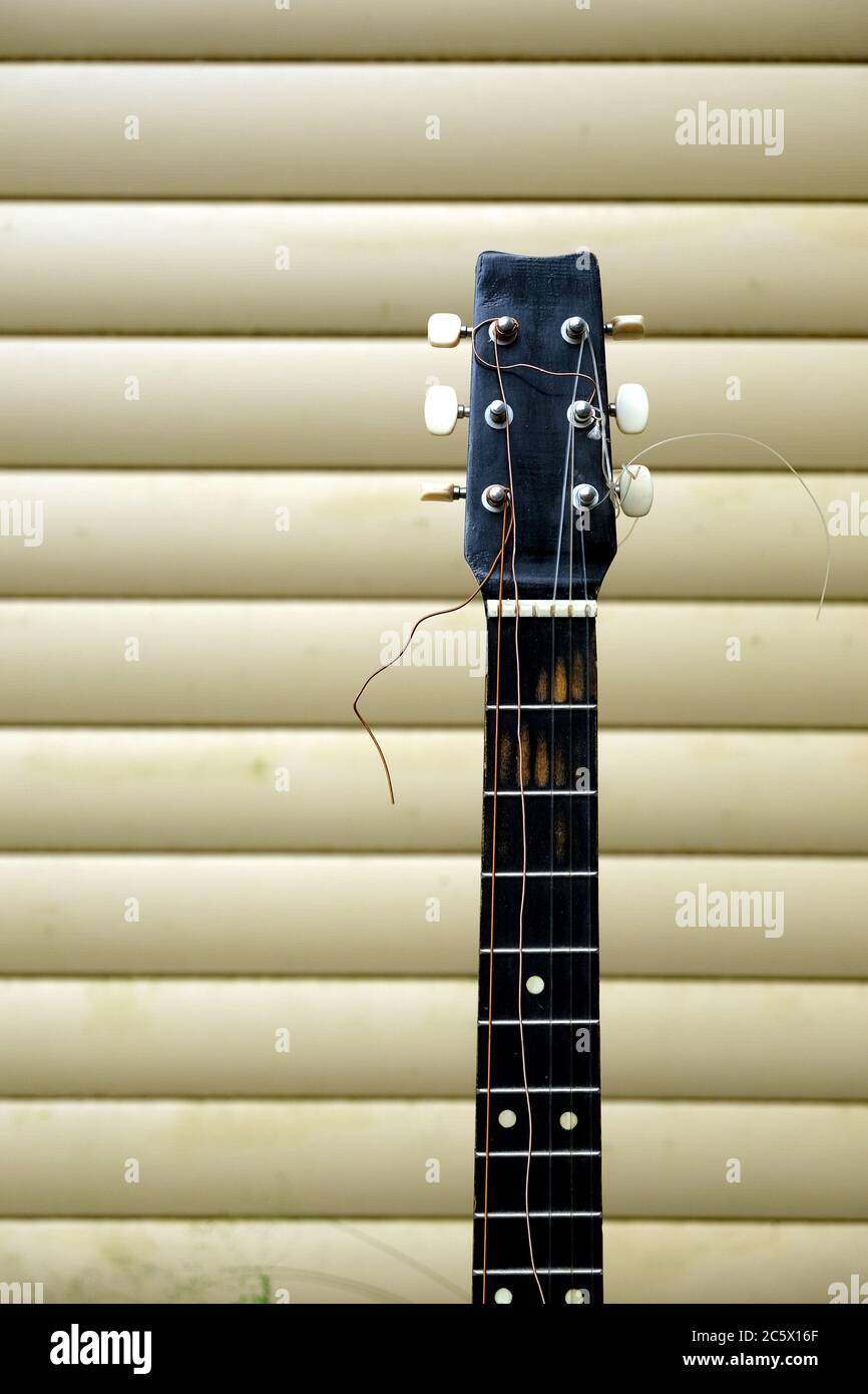 Lungo collo e testa di chitarra in cattive condizioni con corde rotte davanti alla parete della casa vista frontale primo piano Foto Stock