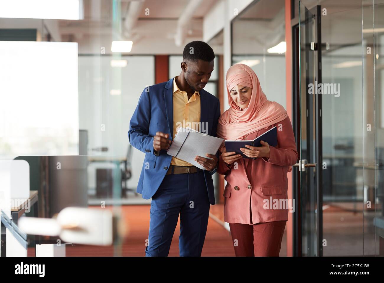 Ritratto di afroamericano che parla con giovane uomo d'affari musulmano e che tiene documenti mentre cammina verso la macchina fotografica nell'interno dell'ufficio, copia spa Foto Stock
