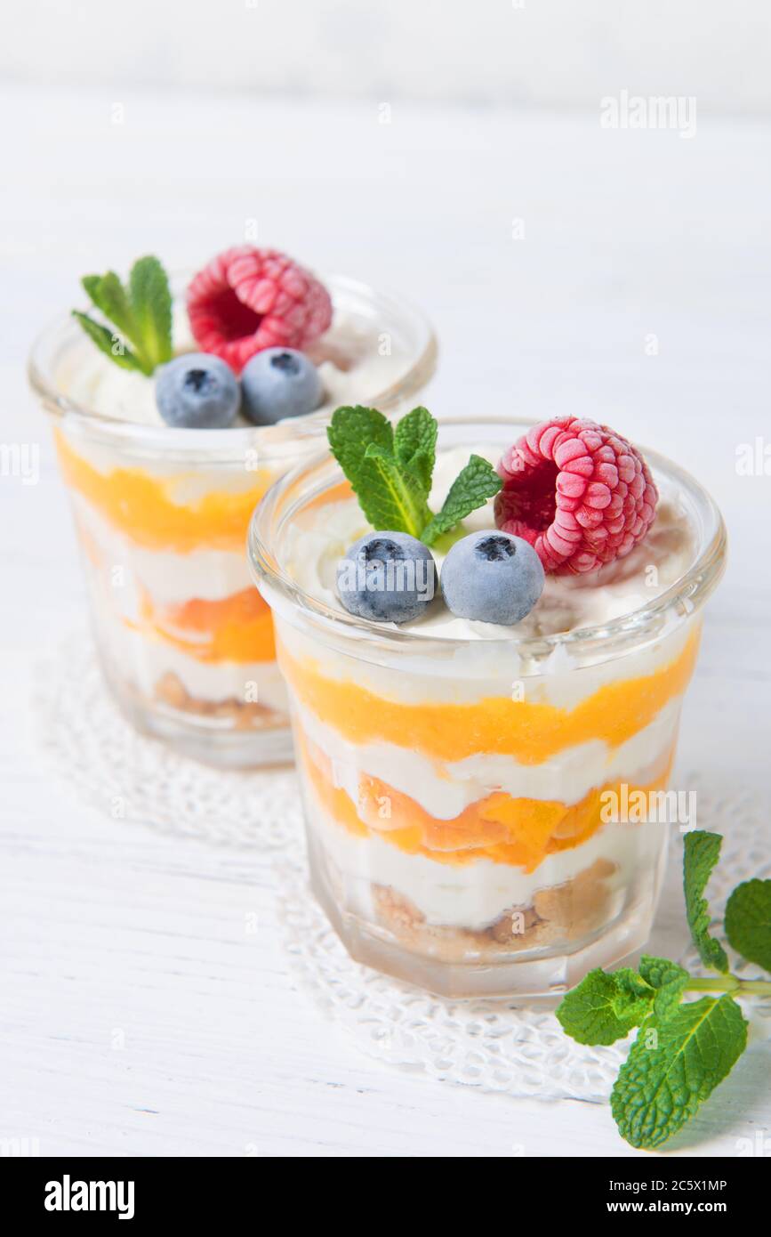 Due porzioni di panna da dessert fatta in casa con pesche, frutti di bosco freschi e menta in un bicchiere su sfondo chiaro Foto Stock