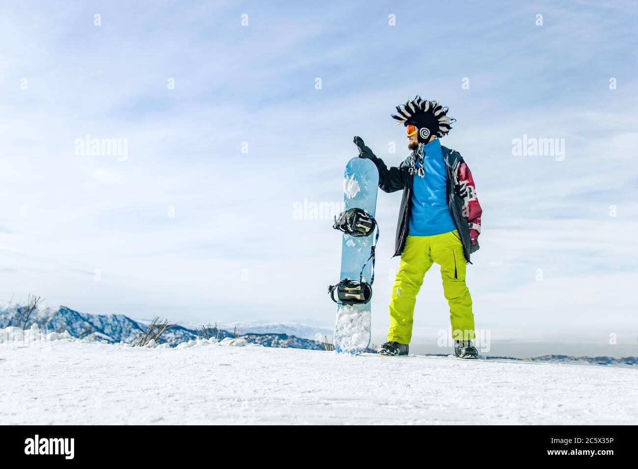 Snowboarder con brandello in maschera da sci con occhiali e un cappello mohawk in pelliccia grande sullo sfondo di un cielo e montagne innevate invernali Foto Stock