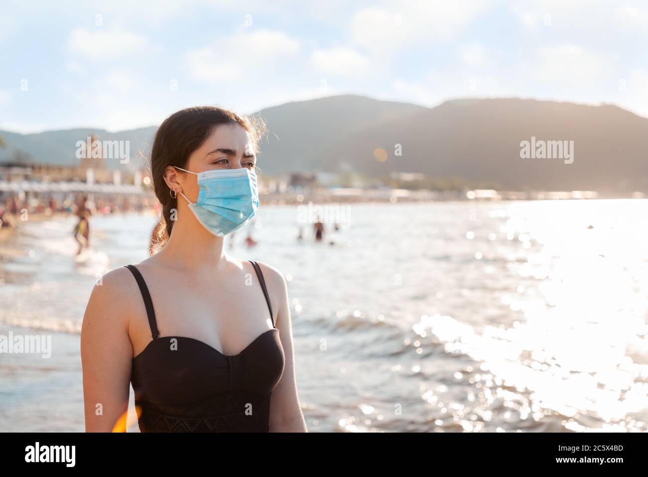 Ritratto di una donna in costume da bagno e maschera medica si trova sulla spiaggia. Concetto di riposo durante una pandemia virale. Spazio di copia. Foto Stock