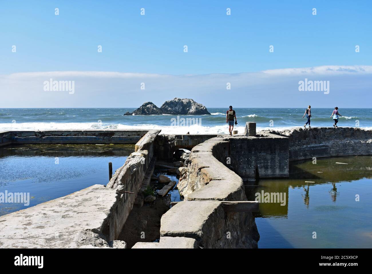 Tre visitatori camminano lungo le rovine dei bagni Sutro a Lands End, San Francisco. L'Oceano Pacifico e le rocce delle foche come sfondo e cielo blu. Foto Stock